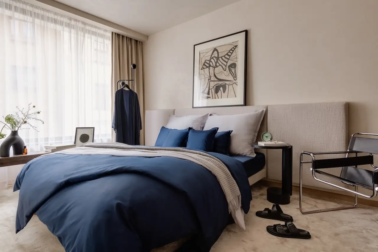 Bett mit dunkelblauer Bettwäsche vor beigefarbener Wand und hellen Vorhängen