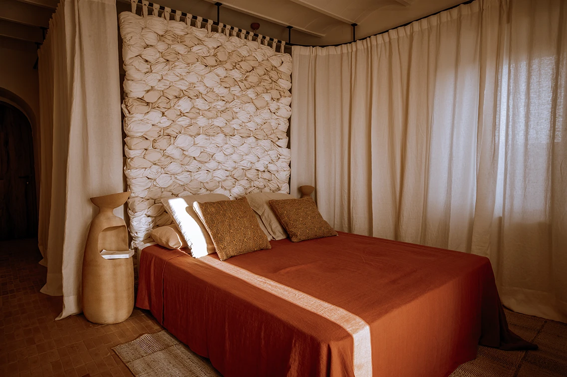 Hotelzimmer im Son Blanc Farmhouse Menorca mit terracottafarbenem Bett, weißem Textil-Kopfteil und warmen Vorhängen im minimalistischen Design.
