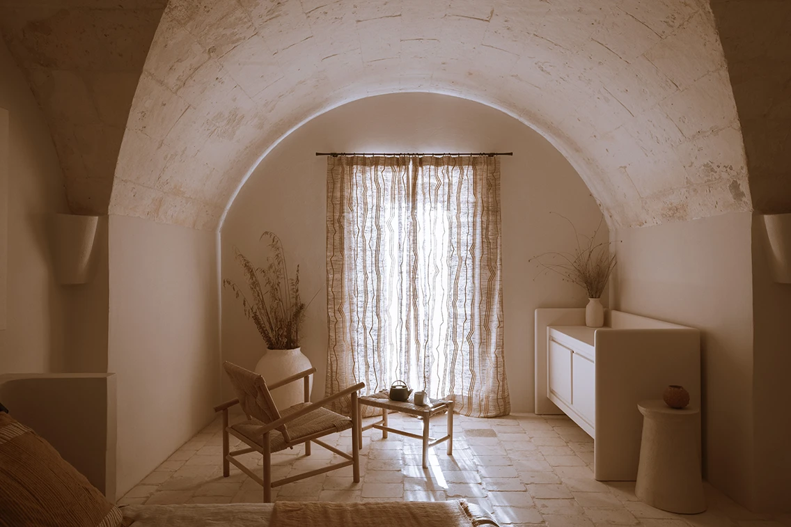 Gemütliche Sitzecke im Gewölbezimmer des Son Blanc Farmhouse Menorca mit Flechtstuhl, Teetischchen und lichtdurchlässigem Vorhang.