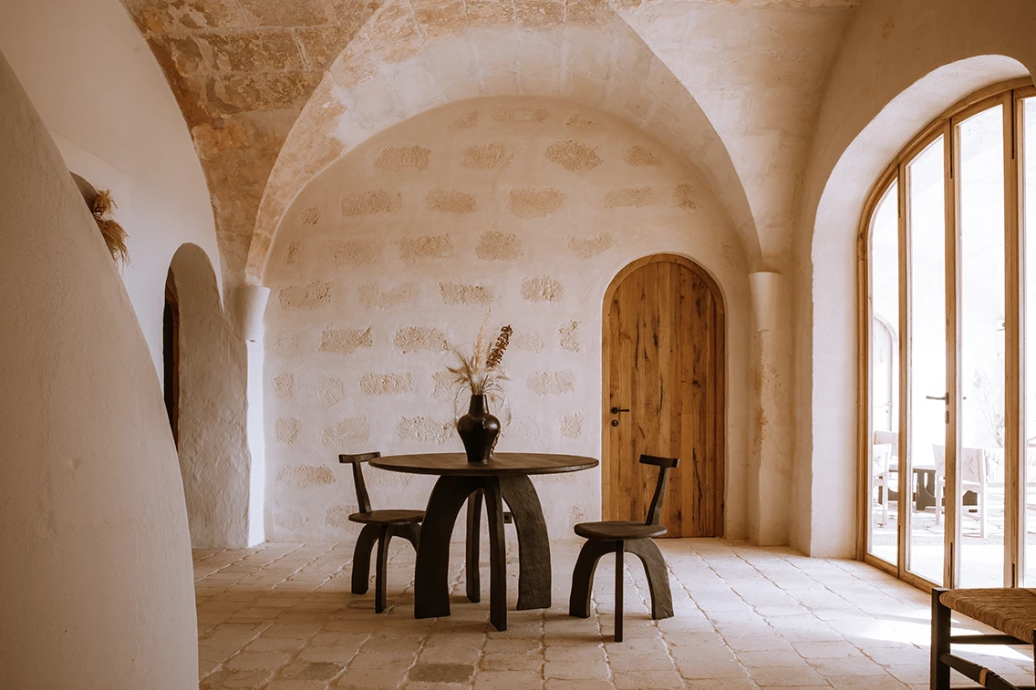 Minimalistischer Eingangsbereich im Son Blanc Farmhouse Menorca mit historischem Kreuzgewölbe, dunklem Holztisch, passenden Designstühlen und rundem Rundbogendurchgang.