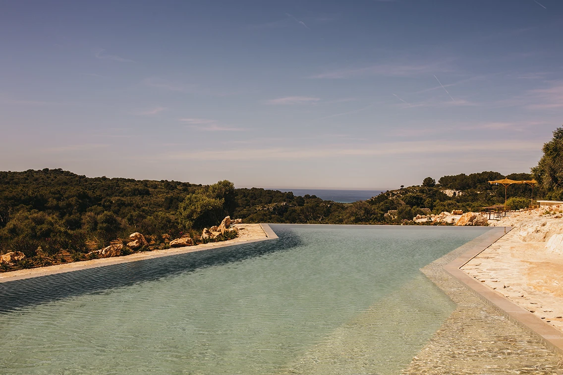 Großzügiger Infinity-Pool im Son Blanc Farmhouse Menorca mit Blick auf die mediterrane Hügellandschaft und das Meer am Horizont.