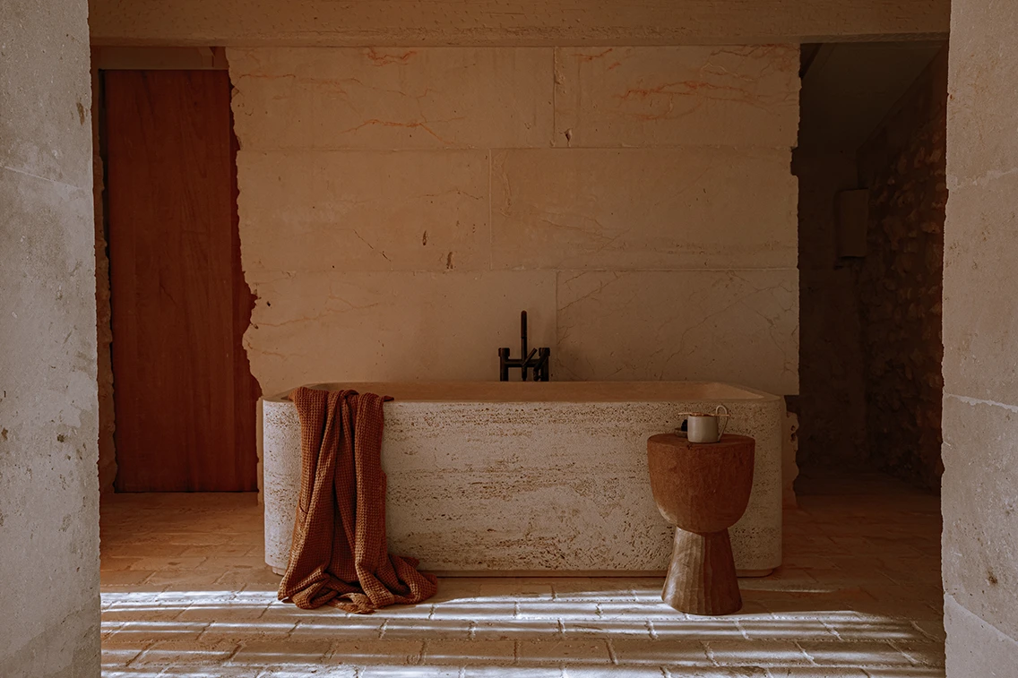 Freistehende Badewanne aus Naturstein mit erdfarbenem Waffelpique-Tuch und Holzbeistelltisch im minimalistischen Badezimmer des Son Blanc Farmhouse.