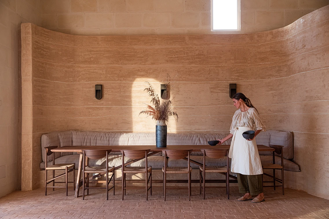 Eleganter Restaurantbereich im Son Blanc Farmhouse Menorca mit langer Holztafel vor einer geschwungenen Stampflehmwand und einer Servicemitarbeiterin beim Eindecken.