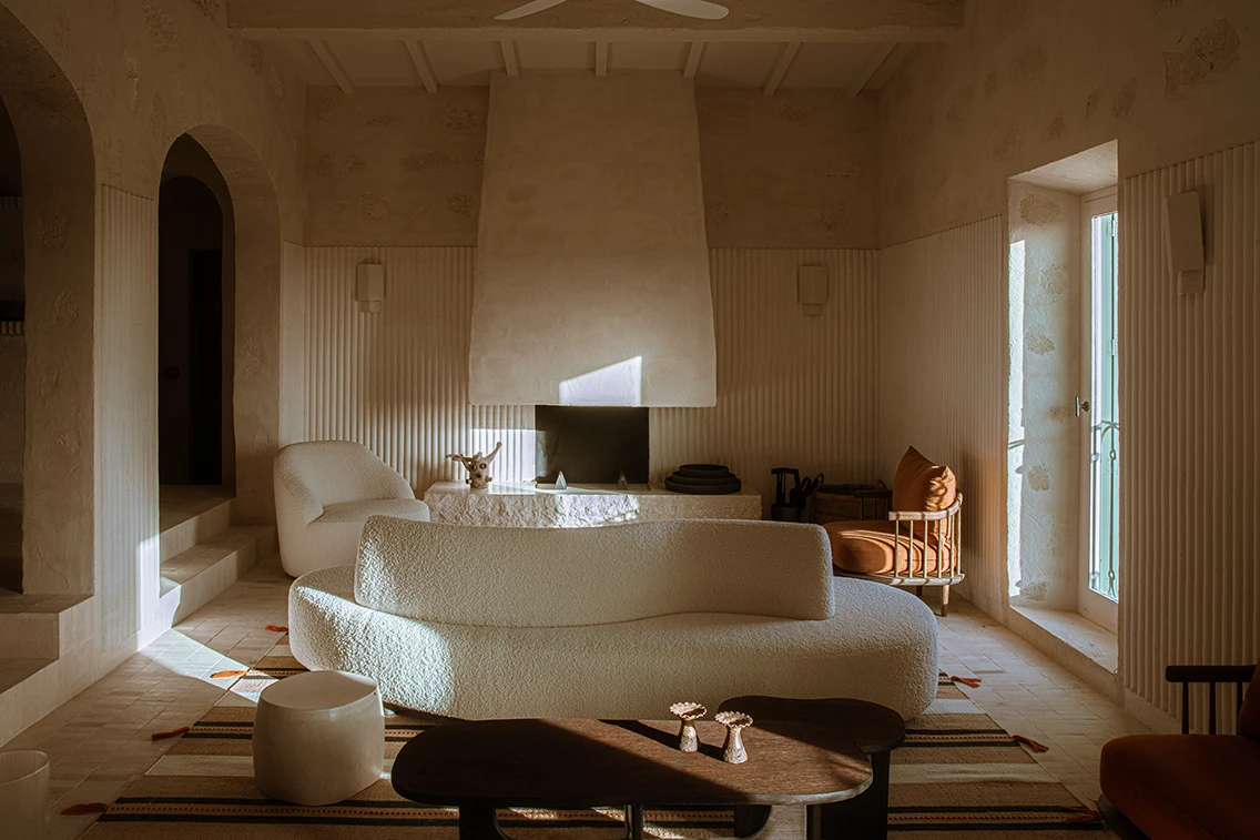 Lichtdurchflutete Kaminlounge im Son Blanc Farmhouse Menorca mit weißem Bouclé-Sofa, erdfarbenem Sessel, Holz-Couchtisch und gewellter Wandverkleidung.