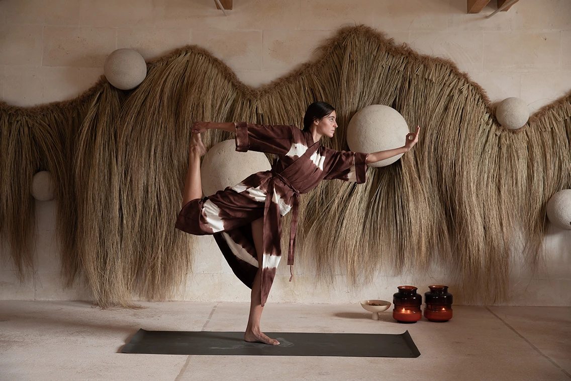 Frau bei einer Yoga-Übung im Yogasaal des Son Blanc Farmhouse Menorca vor einer wandfüllenden Naturfaser-Kunstinstallation und Kerzenlicht.
