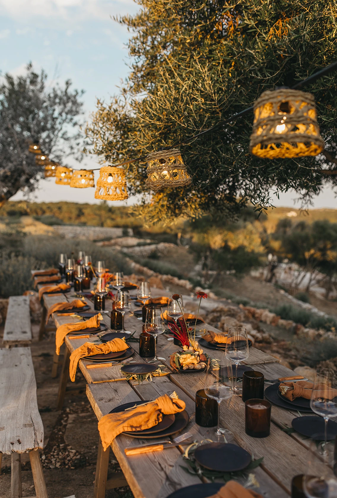 Eingedeckte lange Holztafel für das Communal Dinner im Freien unter Olivenbäumen mit Lichterkette und rustikaler Tischdekoration im Son Blanc Farmhouse.