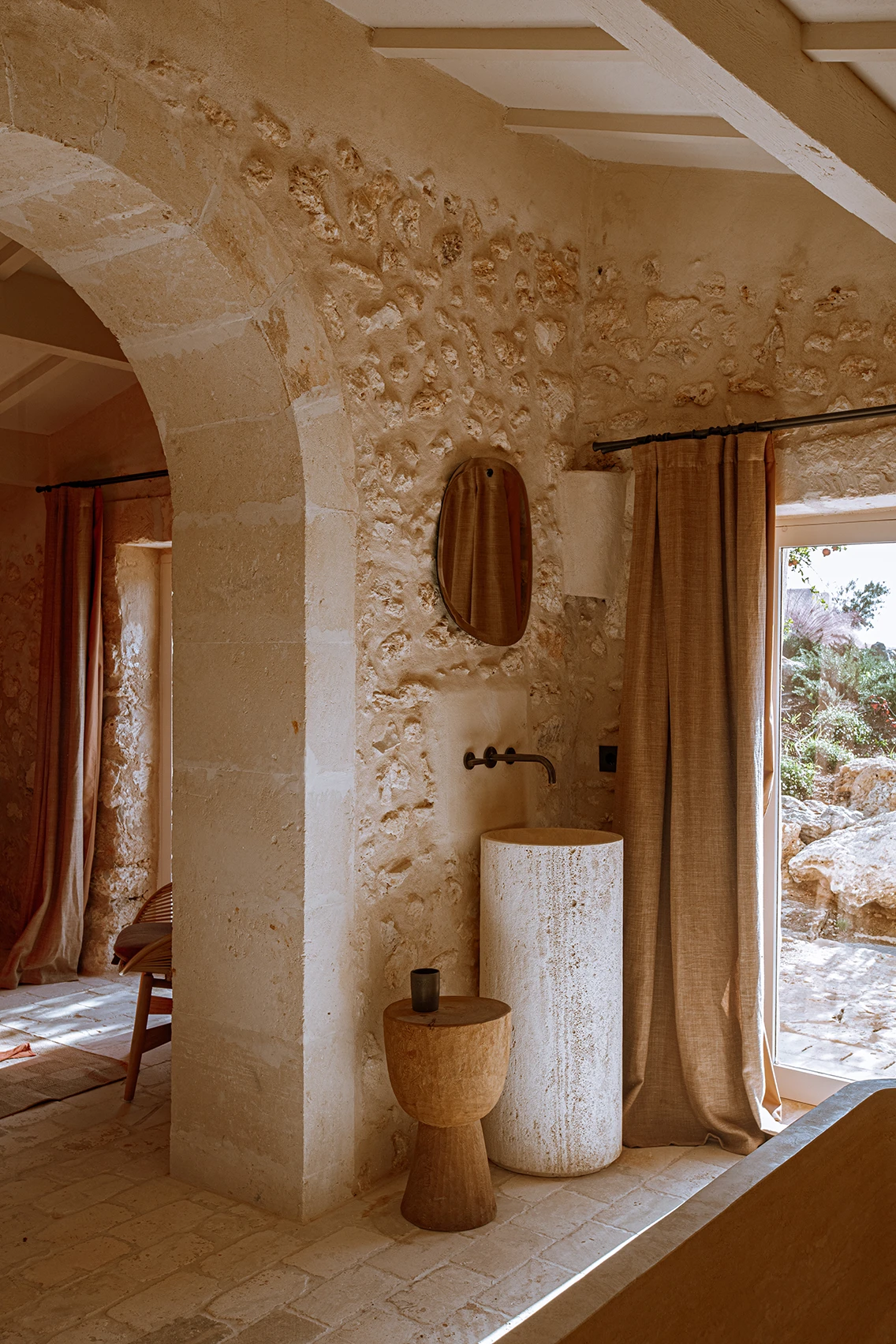 Säulen-Waschtisch aus hellem Naturstein mit wandmontierter Messingarmatur, organischem Spiegel und rustikaler Natursteinwand im Son Blanc Farmhouse.