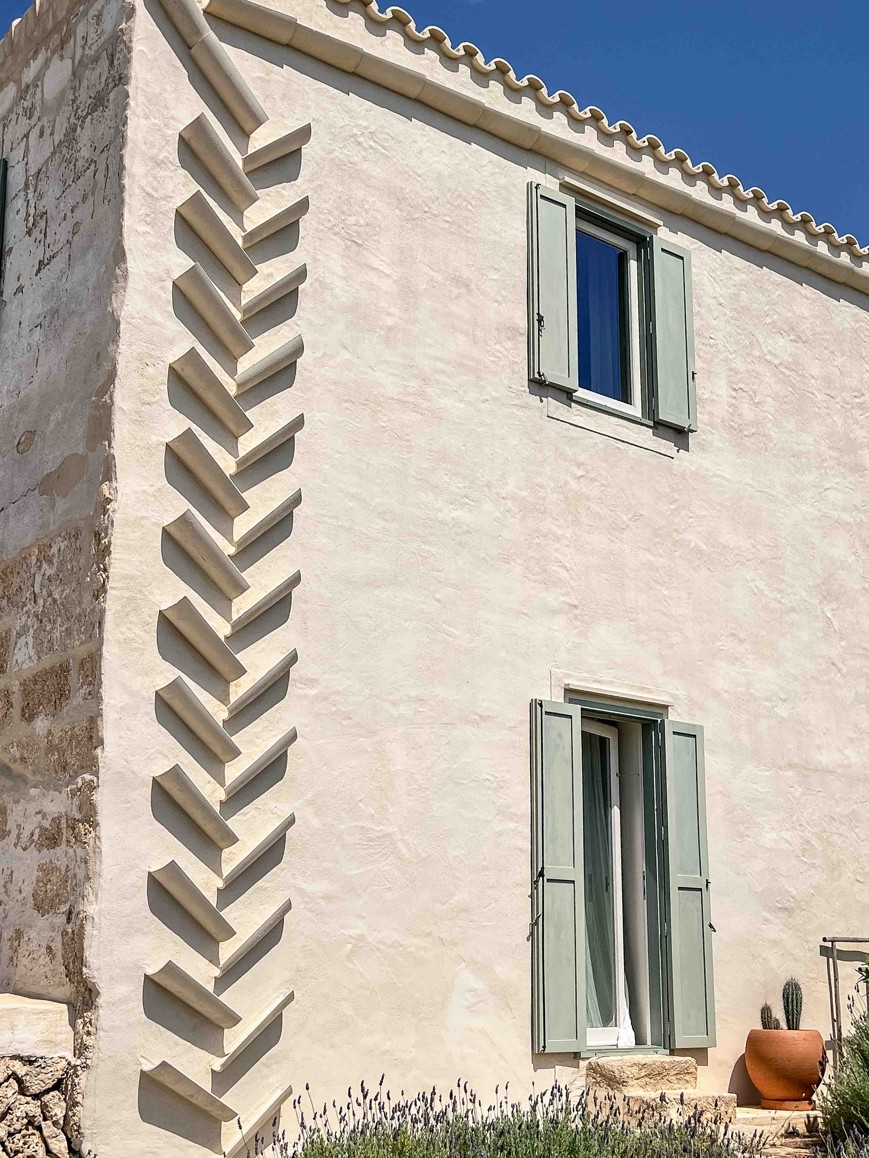 Helle Kalkputz-Fassade des Son Blanc Farmhouse Menorca mit grüngestrichenen Holzläden und dekorativer, zickzackförmiger Ziegel-Regenrinne.