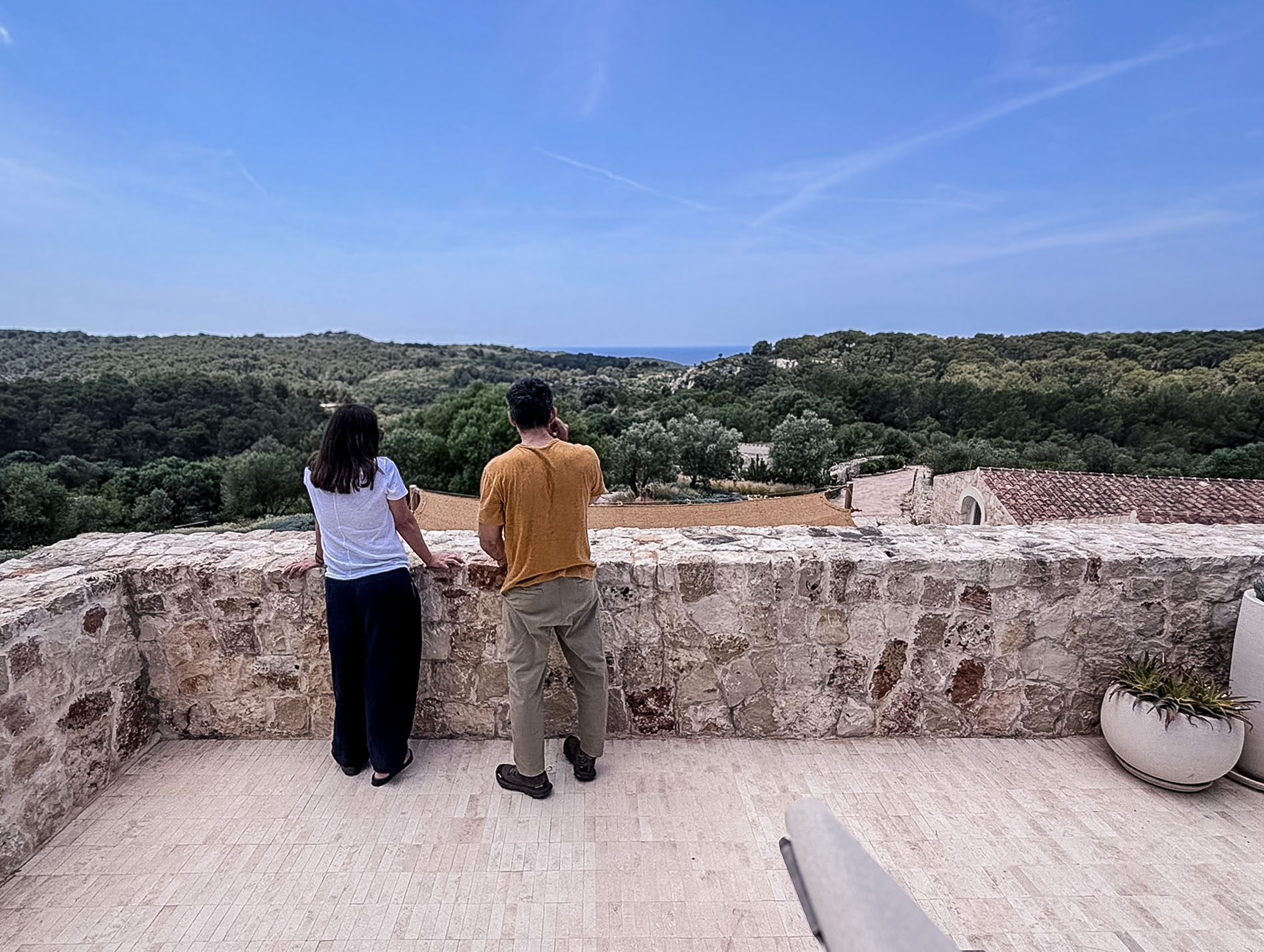 Zwei Personen blicken von der Naturstein-Terrasse des Son Blanc Farmhouse Menorca über die grüne Hügellandschaft bis zum Meer.
