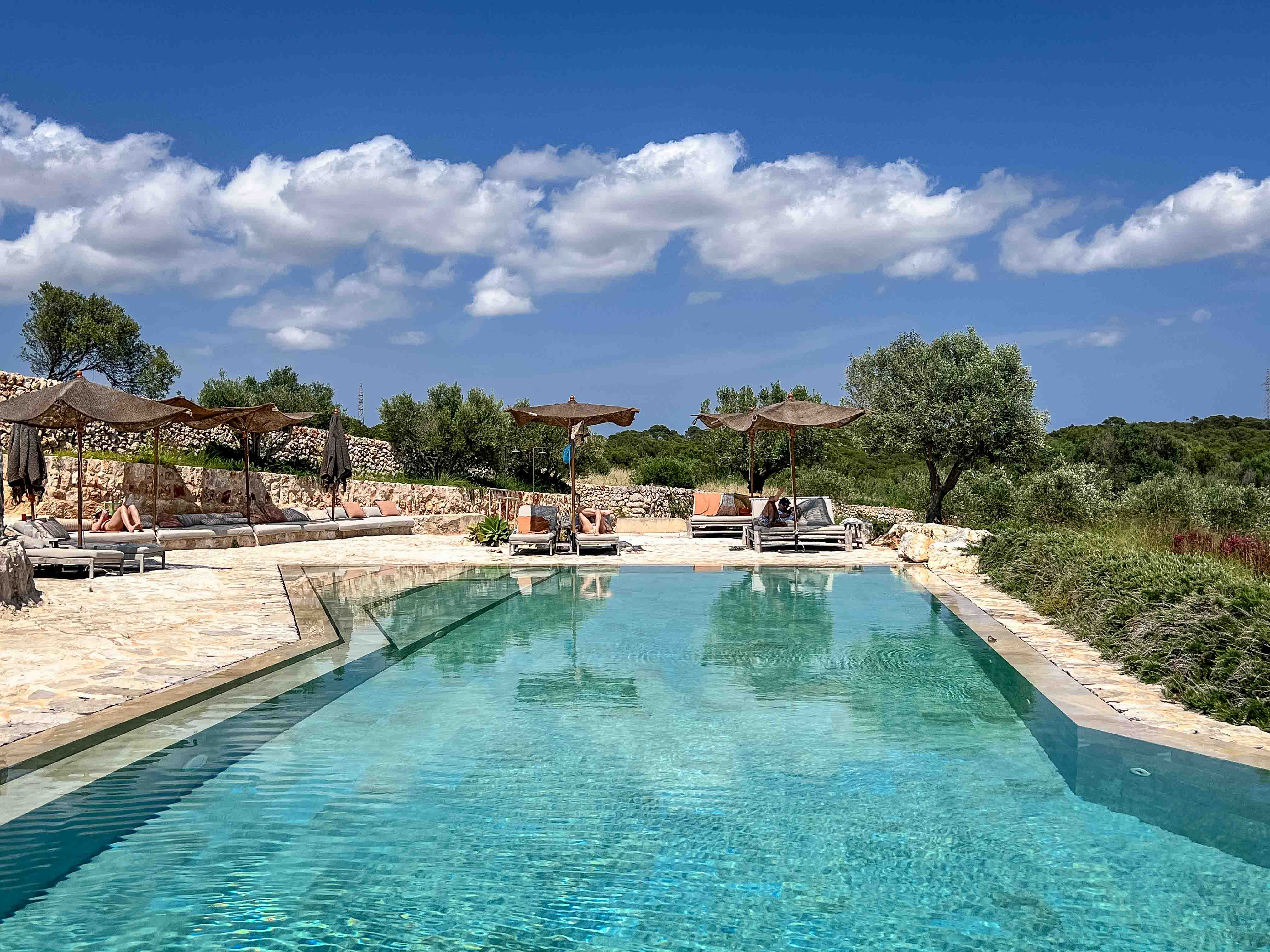 Einbettender Naturstein-Pool im Son Blanc Farmhouse Menorca mit Liegestühlen, Reet-Sonnenschirmen und Blick ins Grüne.