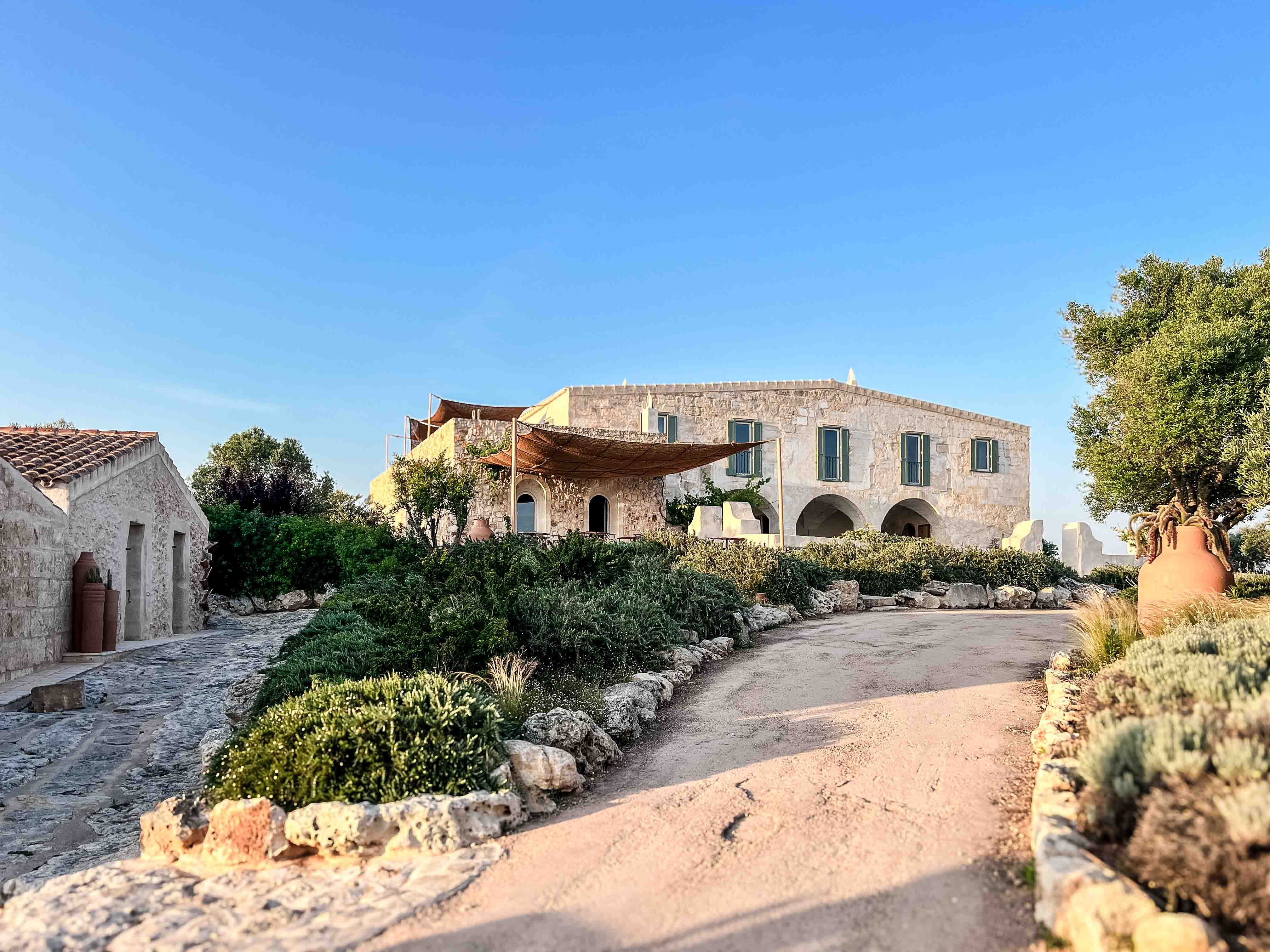 Historisches Finca-Gebäude aus Naturstein im Son Blanc Farmhouse Menorca mit begrüntem Auffahrtsweg und mediterraner Bepflanzung.