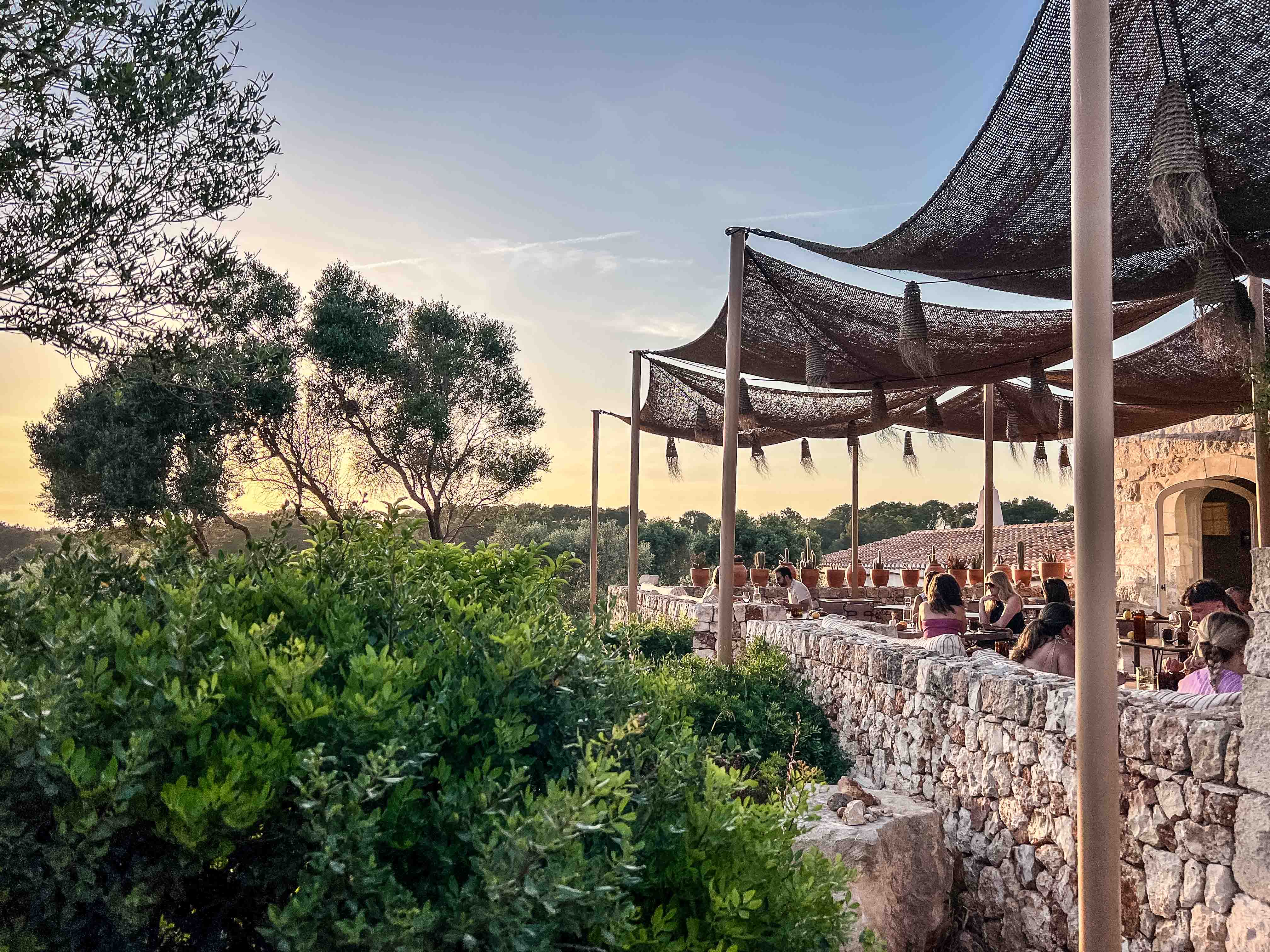 Abendliche Restaurant-Terrasse im Son Blanc Farmhouse Menorca mit Schattendächern, Natursteinmauern und Gästen im warmen Sonnenuntergangslicht.