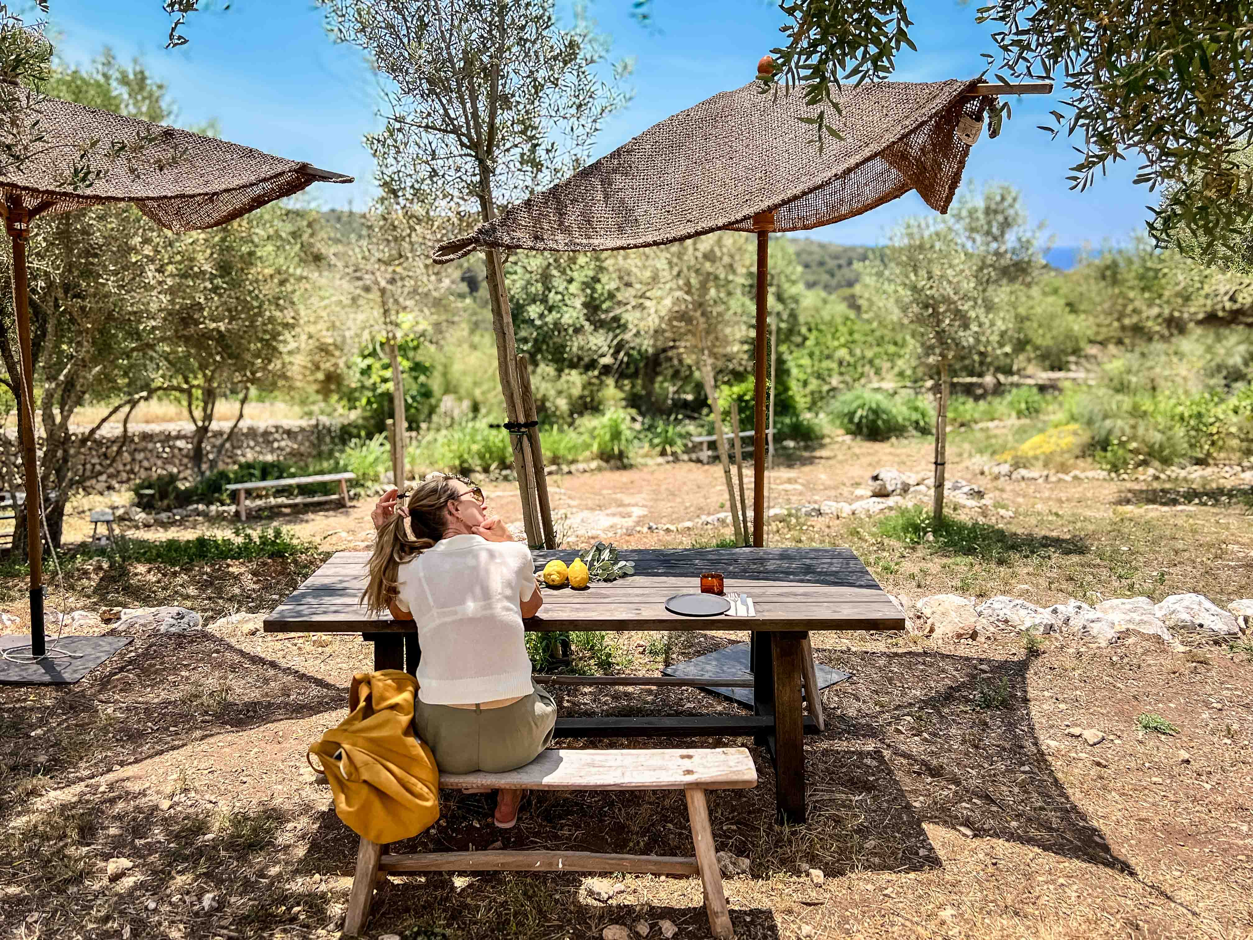 Frau sitzt an einer rustikalen Holzbank unter gewebten Sonnenschirmen im schattigen Garten des Son Blanc Farmhouse Menorca.