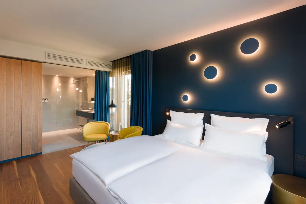 Zimmer EMILU: Blaue Wand, Bett mit weißen Laken, Wandleuchten über dem Bett