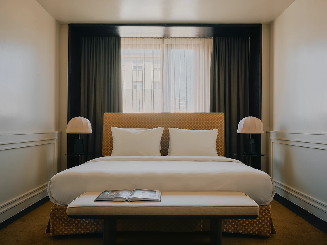 Doppelbett mit gepolstertem Kopfteil, Nachttischlampen und Buch auf der Fußbank in der Junior Suite des Designhotels The Dean München