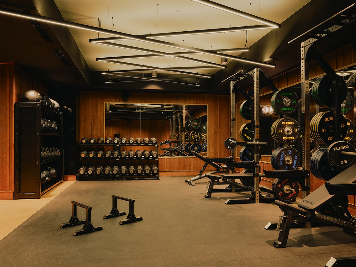 Dunkler Fitnessraum Power Gym mit Kurzhanteln, Eleiko-Gewichtsscheiben und moderner Linear-Deckenbeleuchtung im Hotel The Dean München