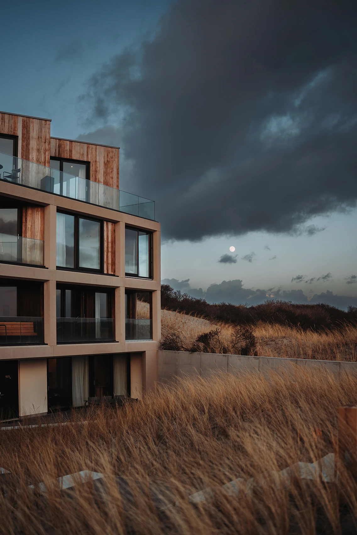 Hotel Elements Terschelling in der Abenddämmerung mit beleuchteten Fenstern, Holz-Beton-Fassade und Vollmond über den Dünengräsern. || © Anne Herens