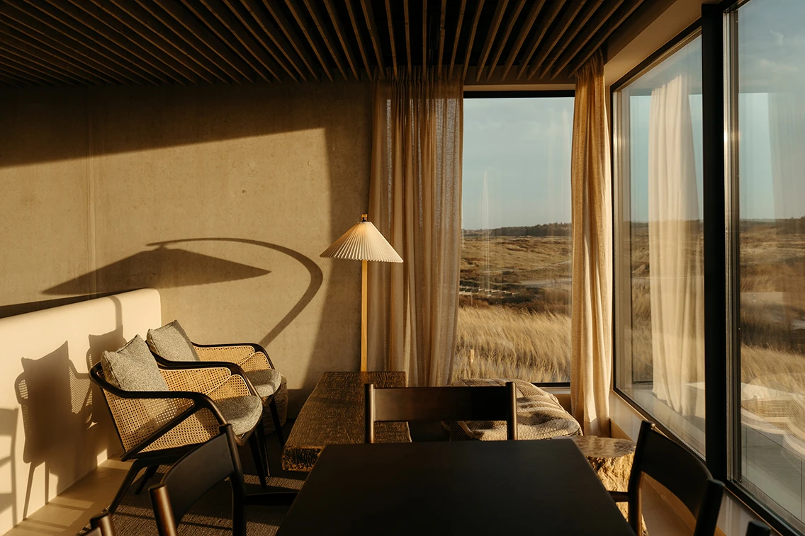 Einladende Sitzecke im Restaurant des Hotel Elements Terschelling mit Wiener-Geflecht-Sesseln, Plissee-Stehleuchte und Panoramablick auf die Dünengräser im goldenen Abendlicht. || © Chantal Arnts