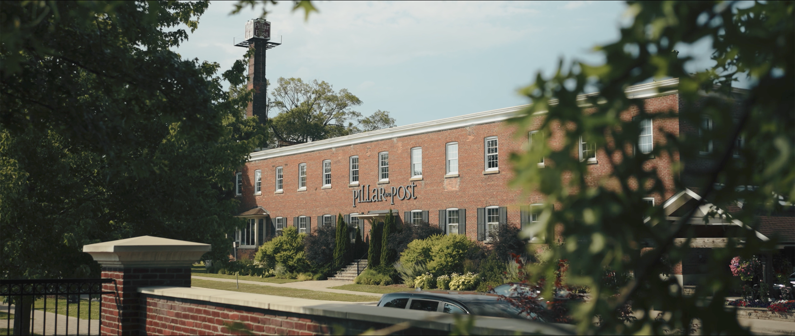 Brick building with a sign reading 'Pillar and Post' partially obscured by trees and a fence in foreground.