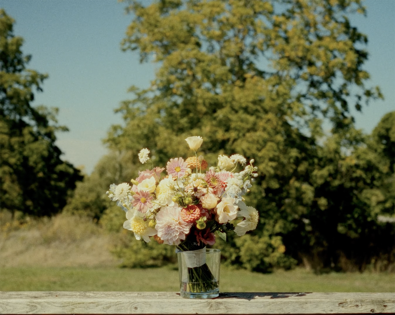 Bouquet of pastel-colored flowers in a glass vase on a wooden surface with green trees in the background.