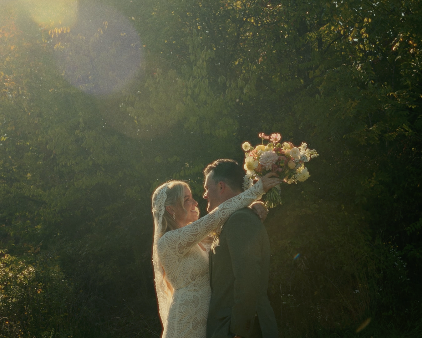 Bride and groom embracing outdoors with bouquet under soft sunlight and green foliage background.