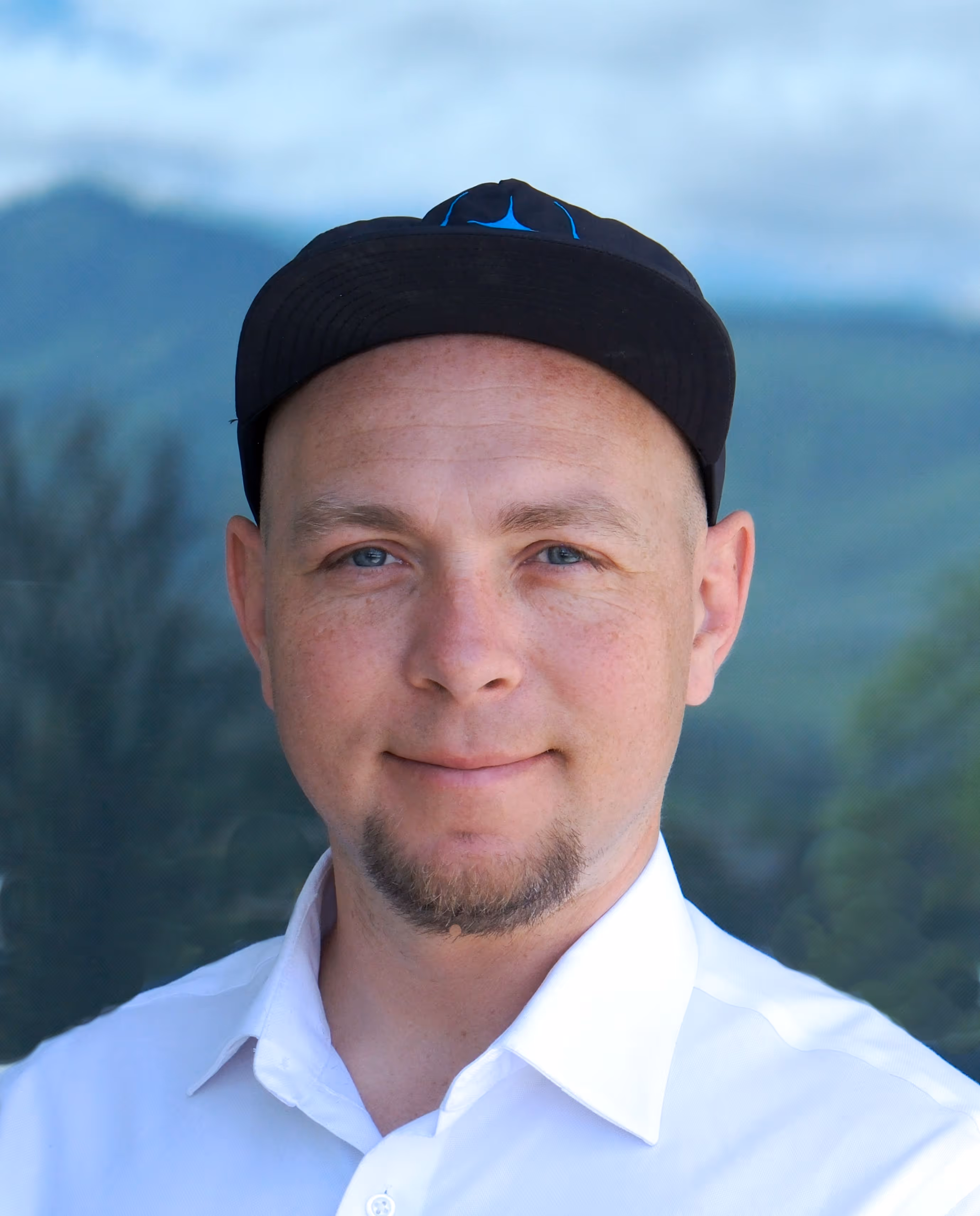 Man with blue eyes and goatee wearing a black cap and white collared shirt, with blurred mountains in the background.