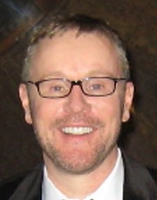 Smiling man with light brown hair, glasses, beard, and suit jacket against a dark background.