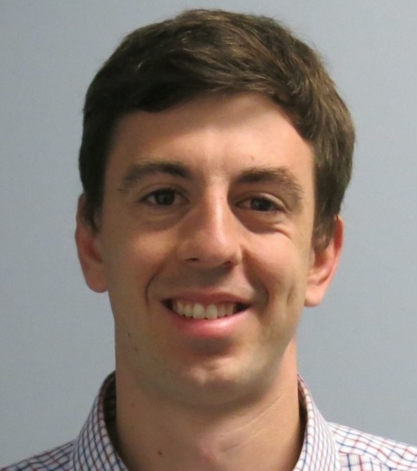 Portrait of a smiling young man with short brown hair wearing a checkered shirt against a plain background.