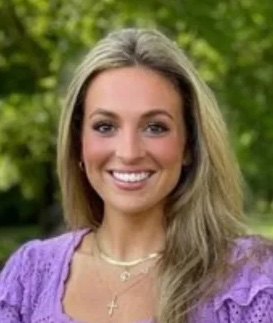 Smiling woman with long blonde hair wearing a lavender top and gold necklaces, standing outdoors with greenery in the background.