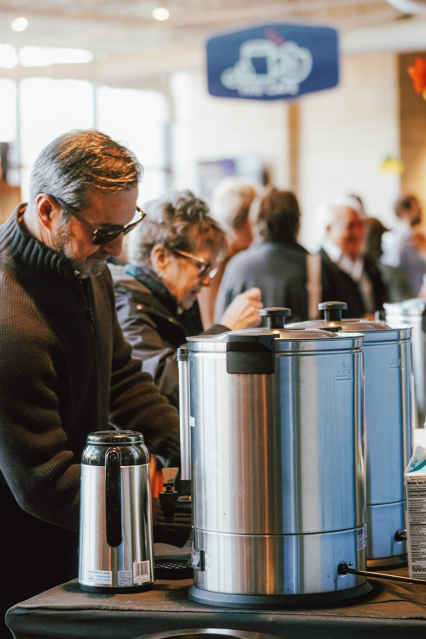 Two people serving themselves coffee from stainless steel beverage dispensers at a counter during a Sunday morning.
