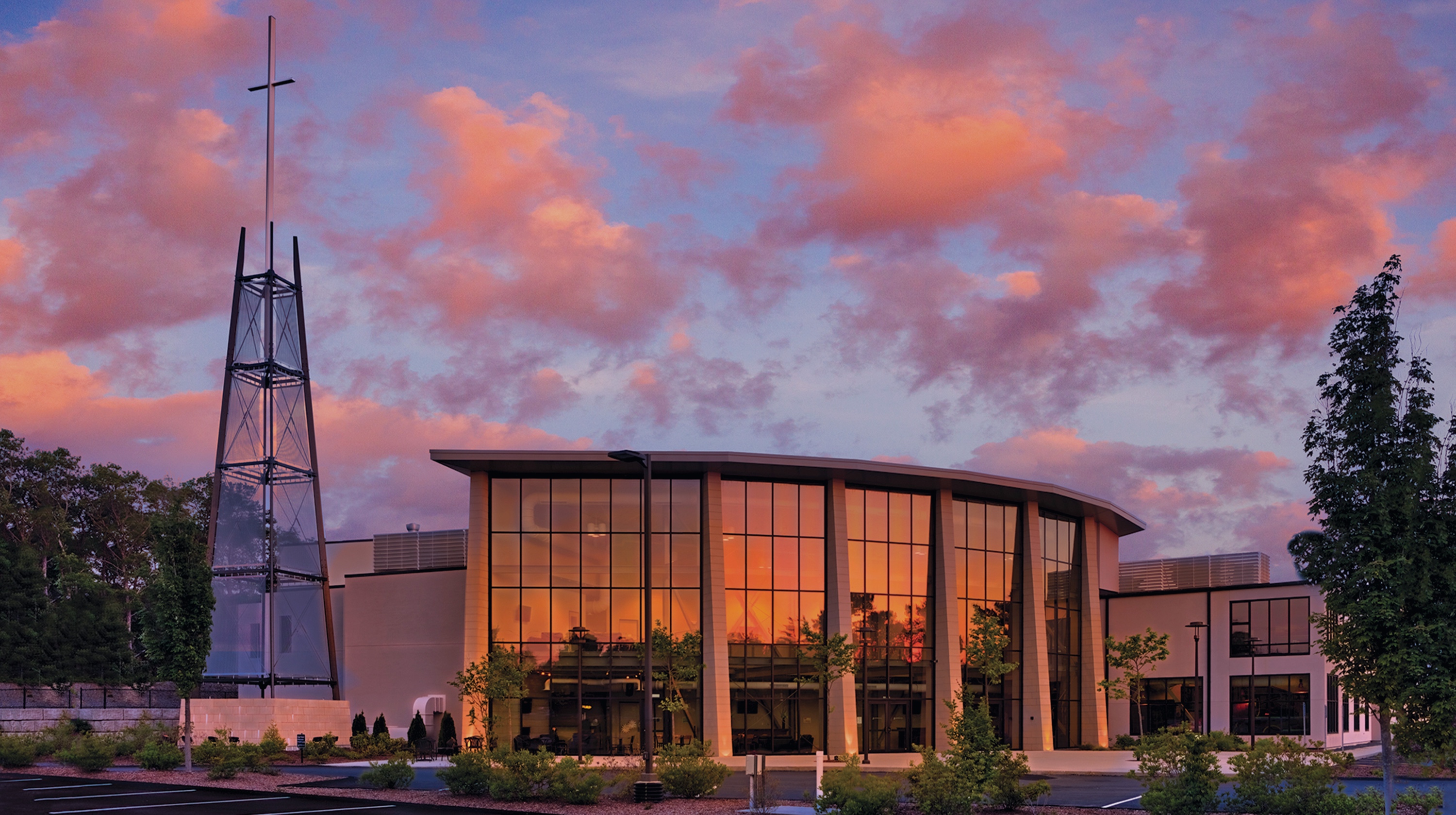 Modern church building with large reflective windows and a tall metal cross tower at sunset with pink and orange clouds.