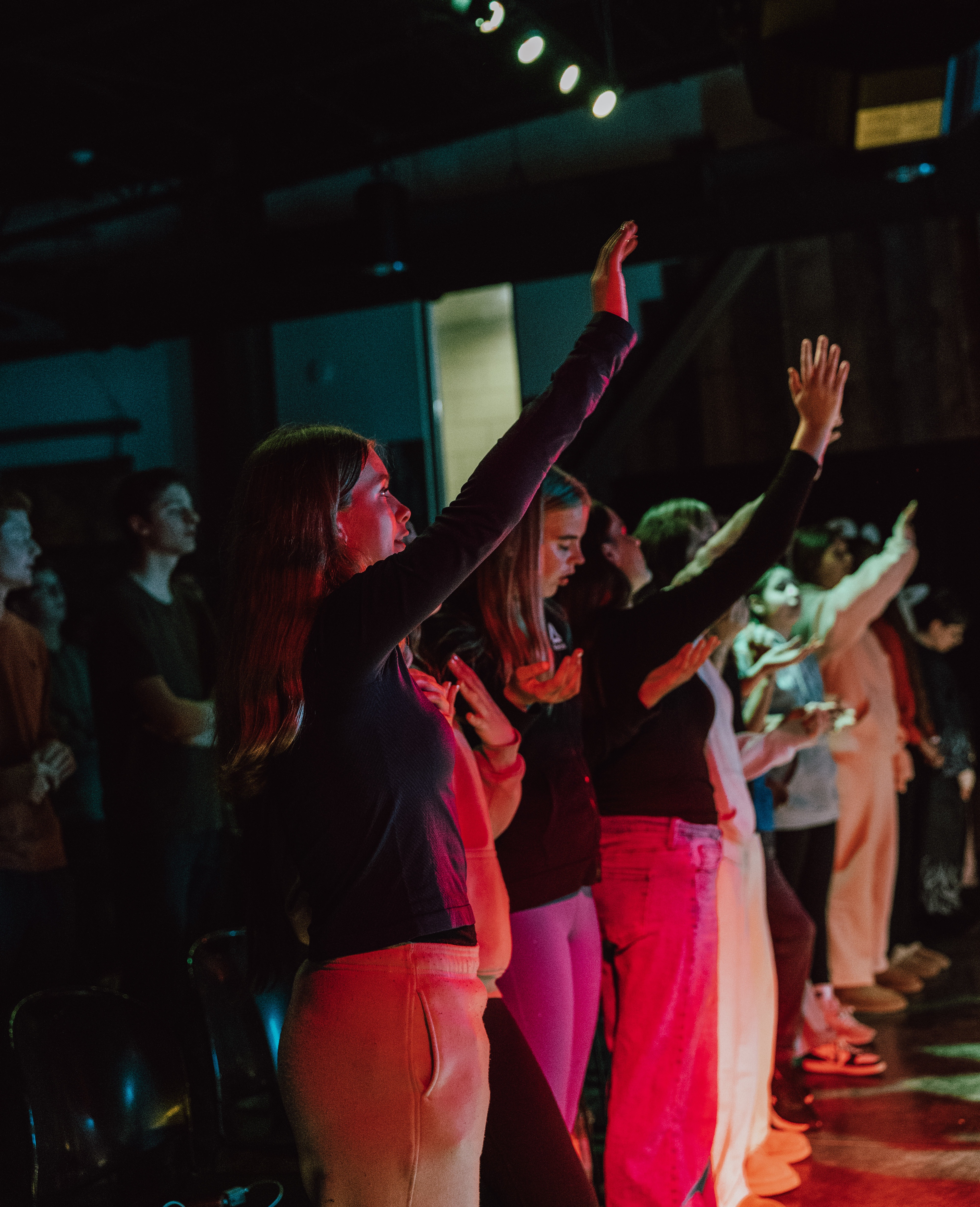 Group of young people standing indoors with some raising their hands in worship, illuminated by colorful stage lighting.