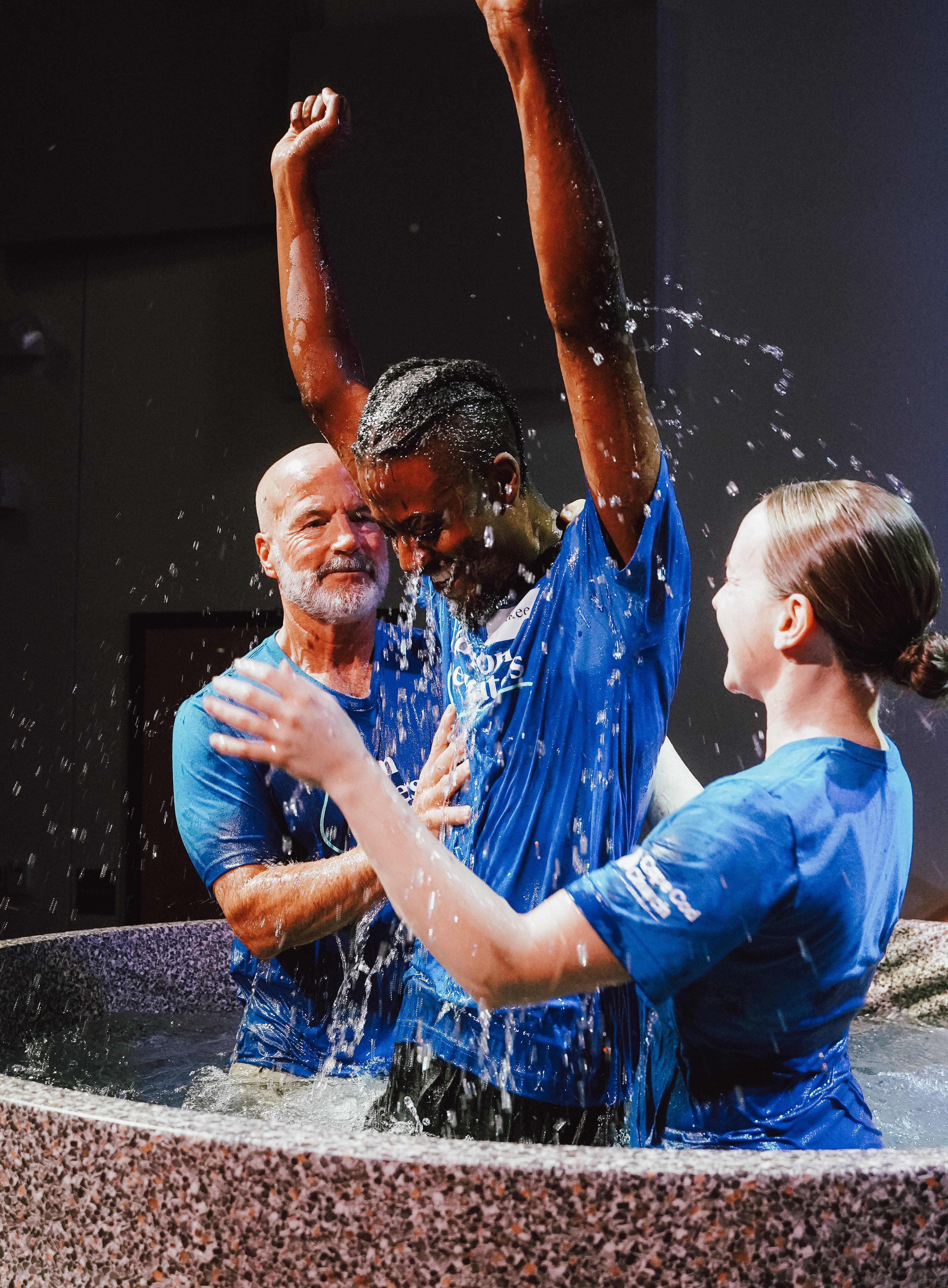 A young man raising his arms in celebration while being baptized with water by two people in blue shirts in a baptismal pool.