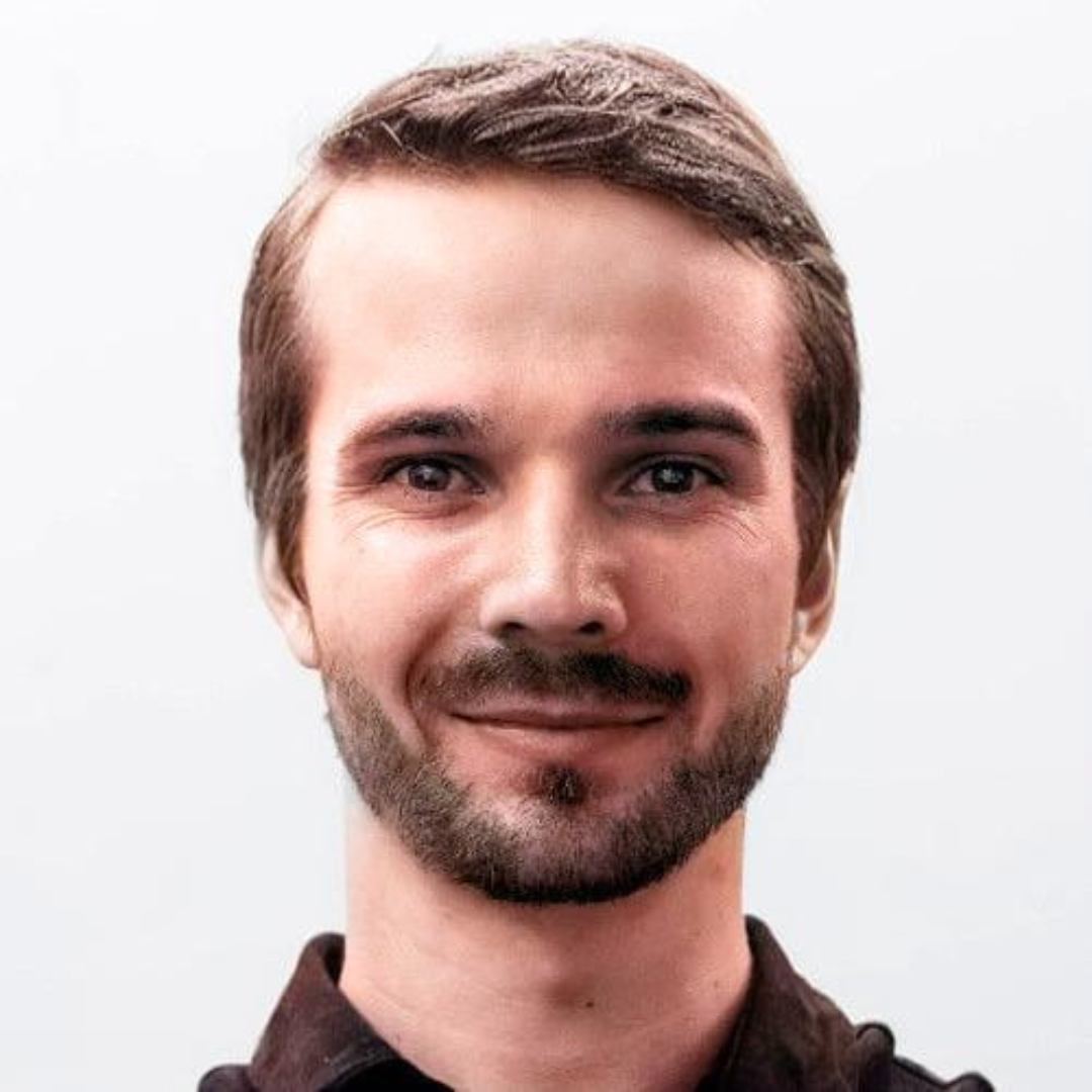 Young man with short brown hair and a beard smiling against a plain light background.