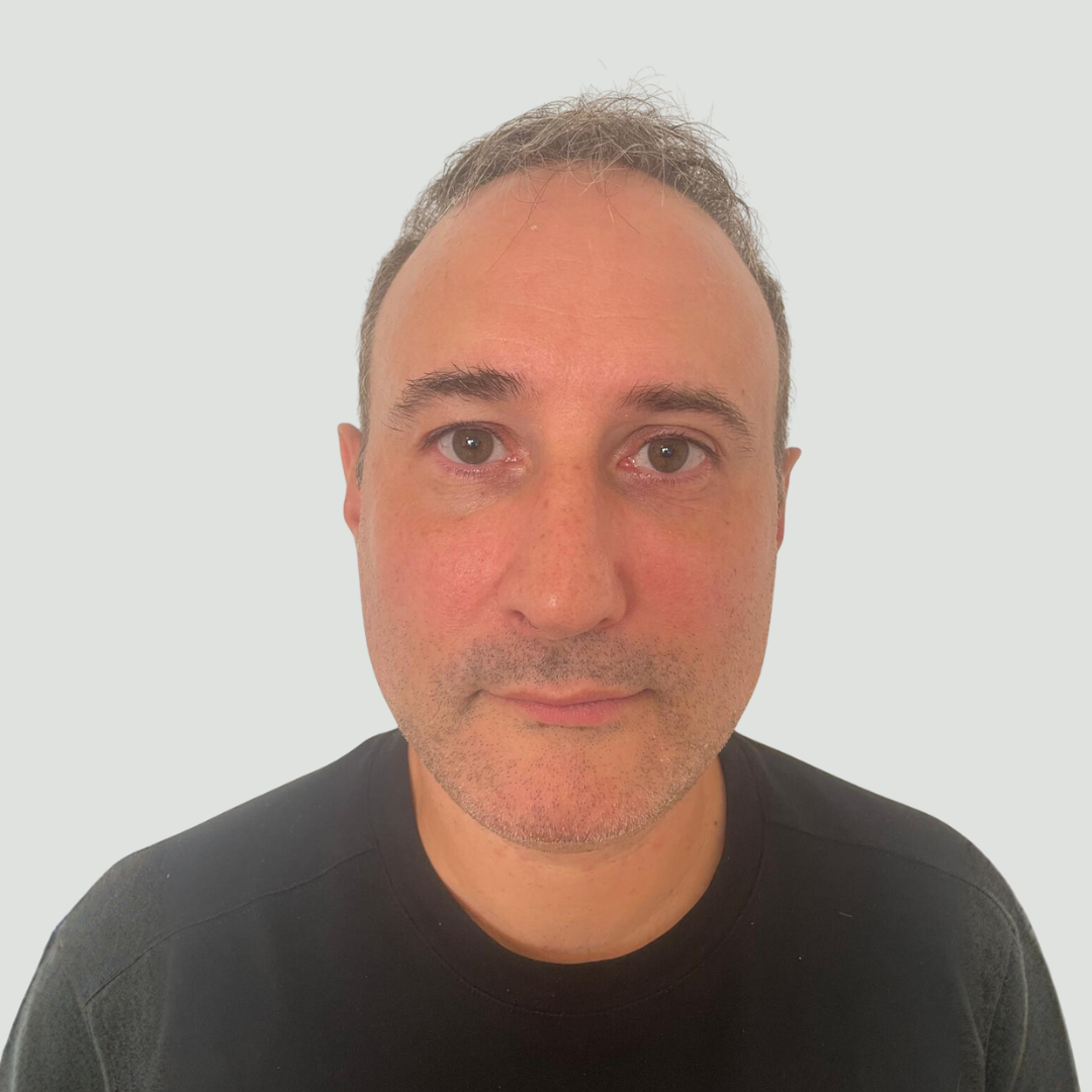 Close-up portrait of a middle-aged man with short dark hair and wearing a black shirt against a light gray background.