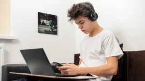 young man wearing headphones and using his phone while having a laptop in front of him, there is a photo tile on the wall behind him