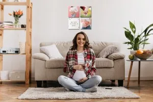 pregnant young woman sitting on a rug on the floor in front of a couch with foam photo tiles on the wall behind it