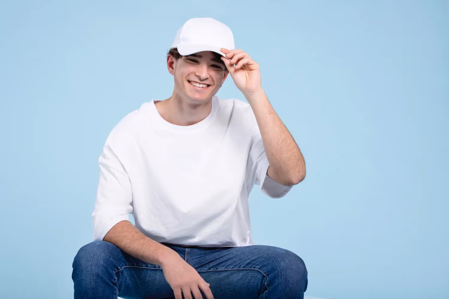 Smiling young man wearing a plain white trucker hat and casual white t-shirt, sitting against a light blue background — ideal lifestyle shot for POD hat mockup or streetwear brand imagery.