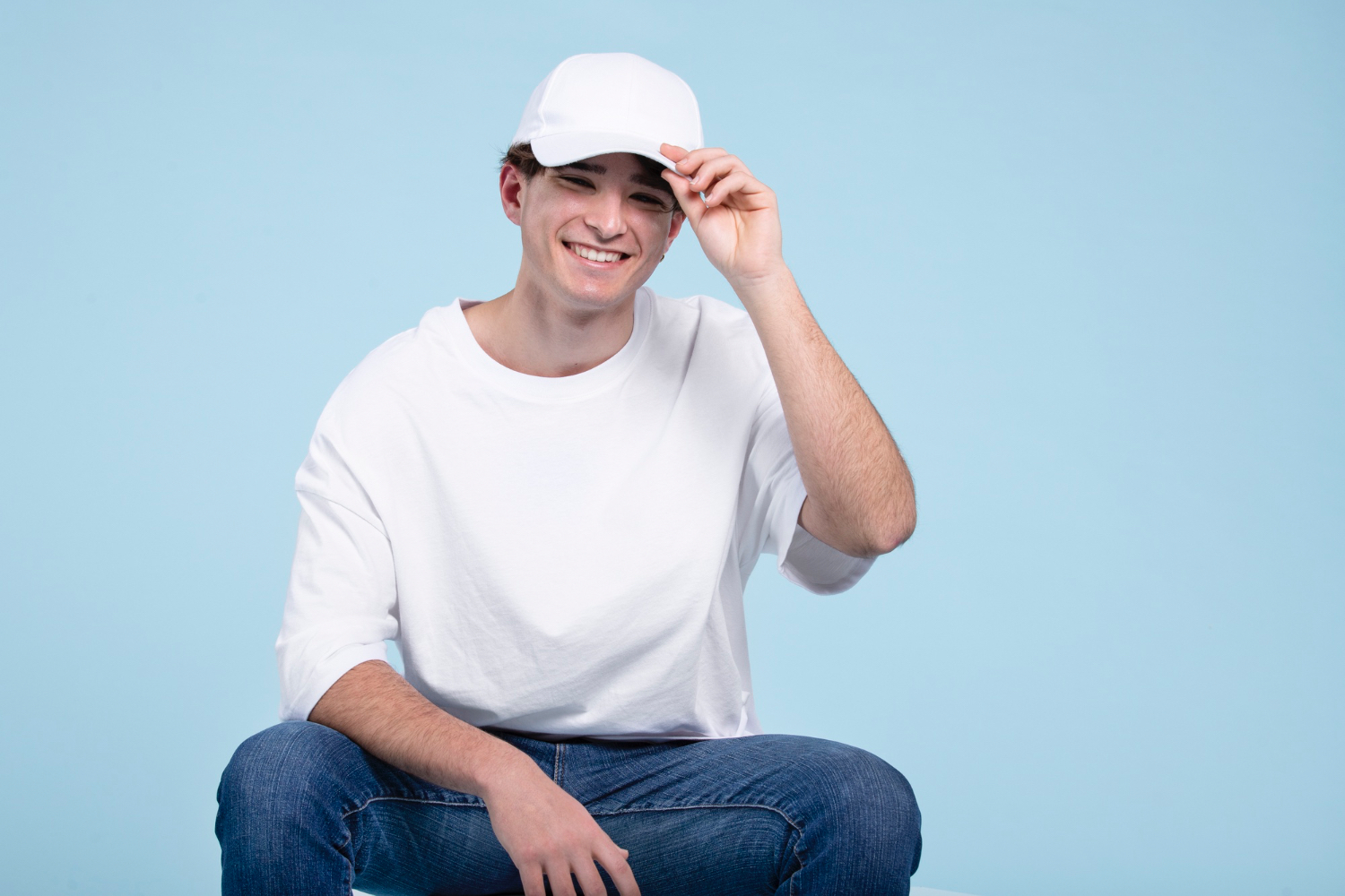 Smiling young man wearing a plain white trucker hat and casual white t-shirt, sitting against a light blue background — ideal lifestyle shot for POD hat mockup or streetwear brand imagery.