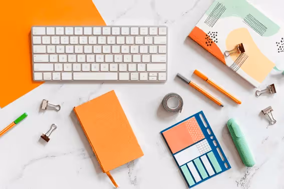 Desk flat lay with orange and pastel-colored stationery, notebooks, pens, sticky notes, and a white computer keyboard on marble surface.
