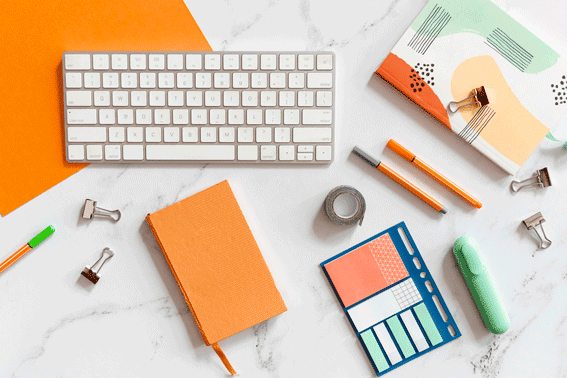 Desk flat lay with orange and pastel-colored stationery, notebooks, pens, sticky notes, and a white computer keyboard on marble surface.
