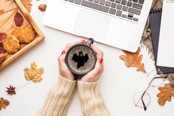 Hands holding coffee mug with bat latte art on autumn desk with cookies, laptop, and fall leaves.
