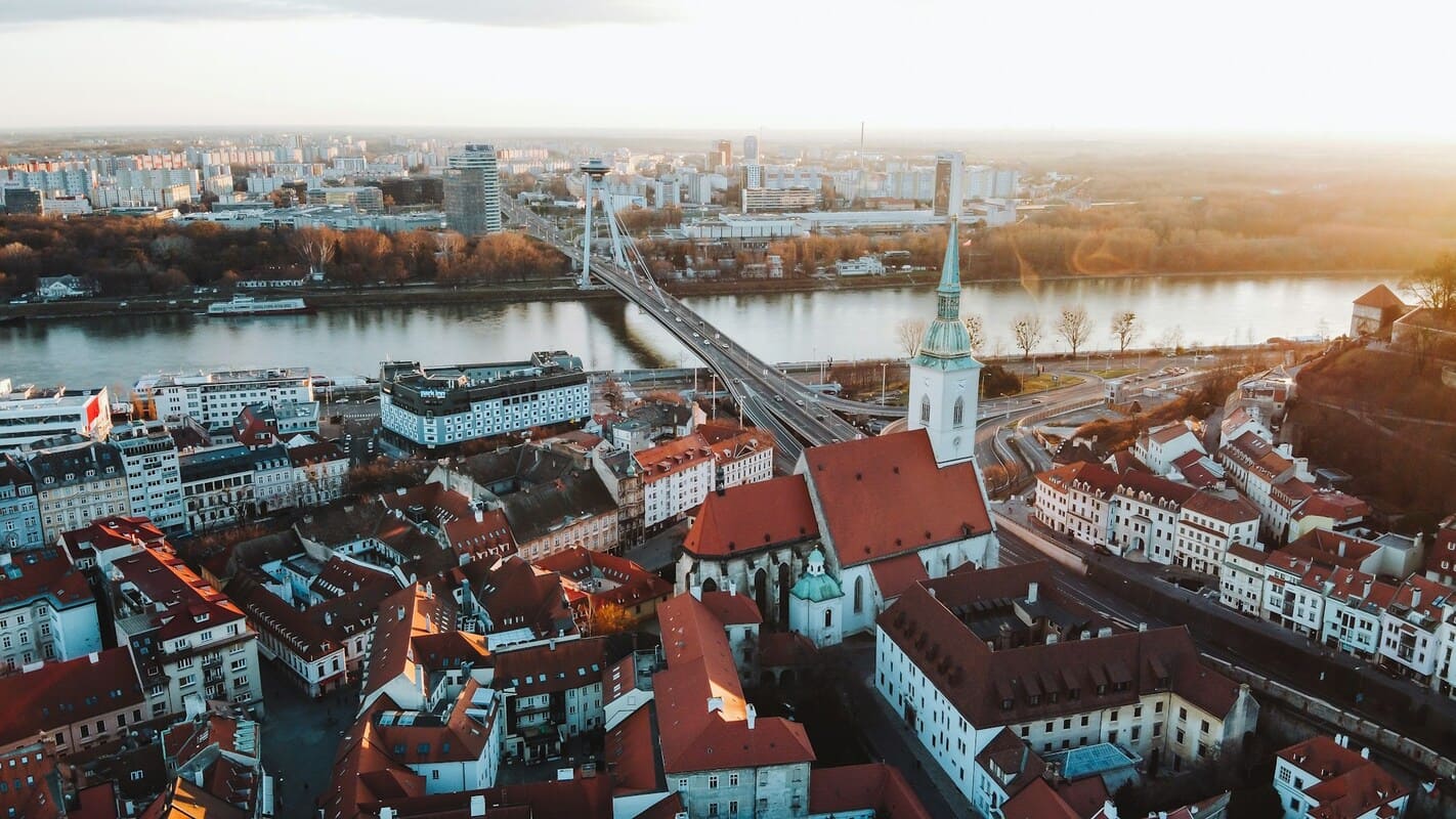 Aerial view of a city with red-roofed buildings, a large church with a tall spire, a river, and a suspension bridge crossing the river at sunset.