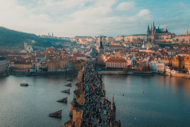 Aerial view of Charles Bridge crowded with people spanning the Vltava River in Prague, with historic buildings and Prague Castle in the background under a cloudy sky.