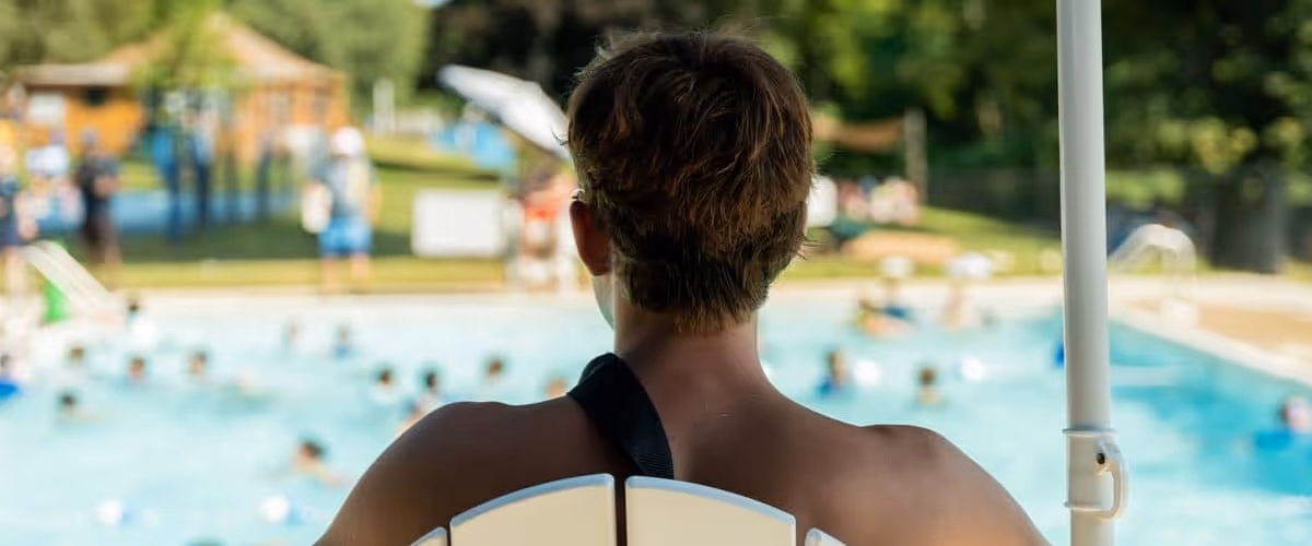 Back view of a lifeguard sitting on a high chair overlooking a busy outdoor swimming pool.