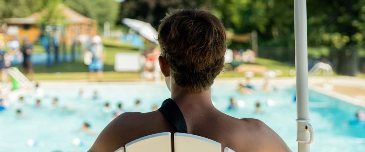 Back view of a lifeguard sitting on a high chair overlooking a busy outdoor swimming pool.