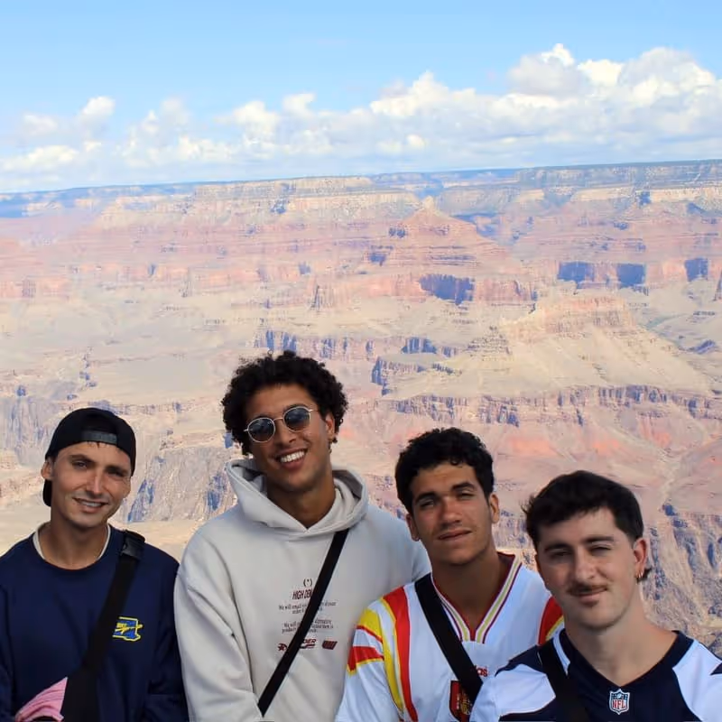 Four young men smiling and posing with the Grand Canyon in the background under a partly cloudy sky.