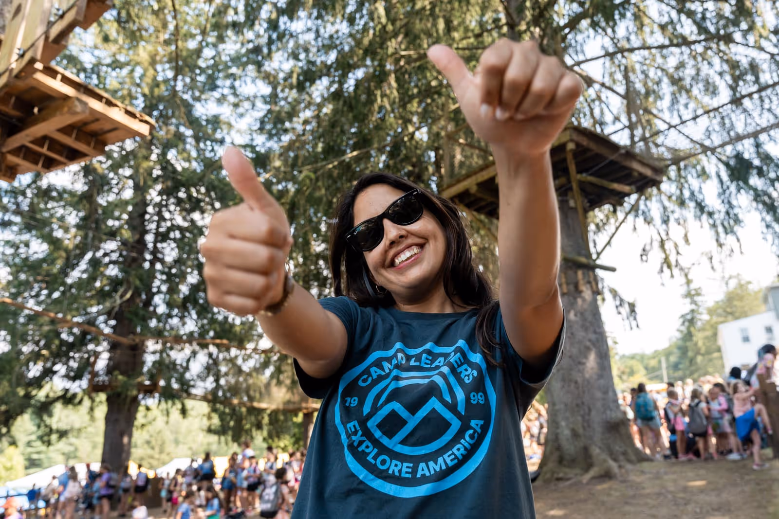 Smiling woman wearing sunglasses and a Camp Leaders Explore America t-shirt giving two thumbs up outdoors at a busy camp with trees and people in the background.