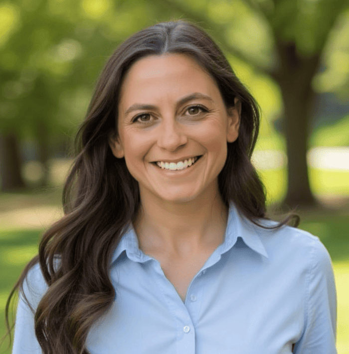 Smiling woman with long brown hair wearing a light blue shirt outdoors with blurred green trees in the background.