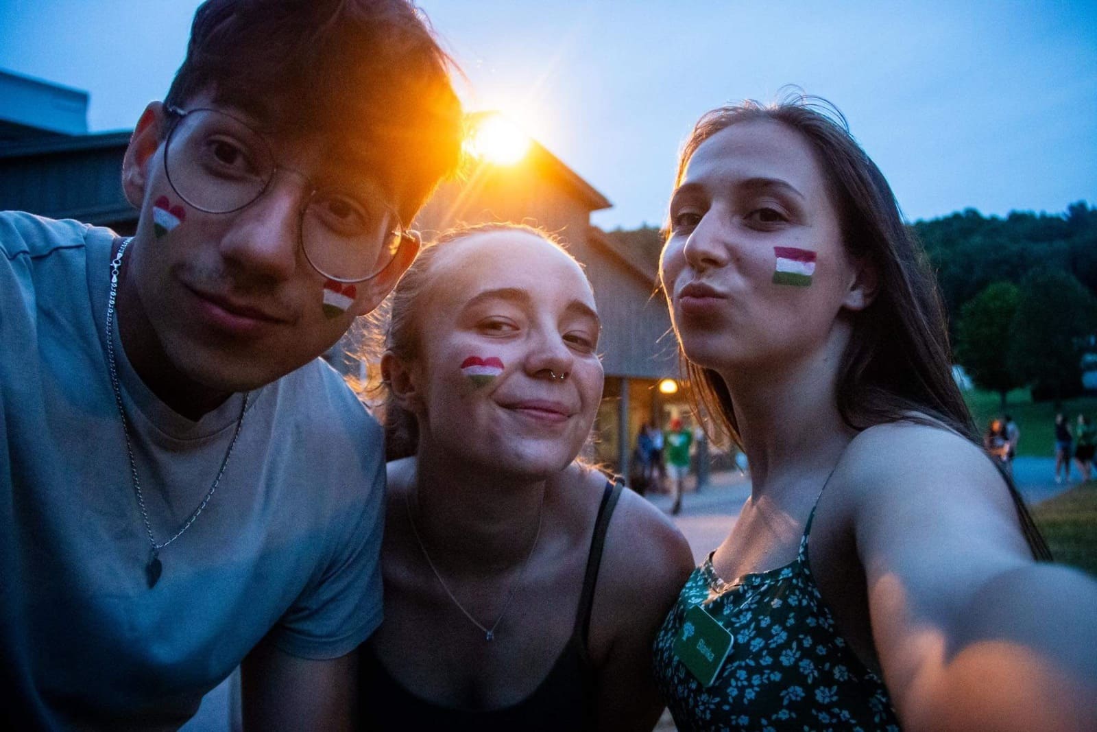 Three young people with face paint of the Hungarian flag posing for a selfie outdoors at dusk.
